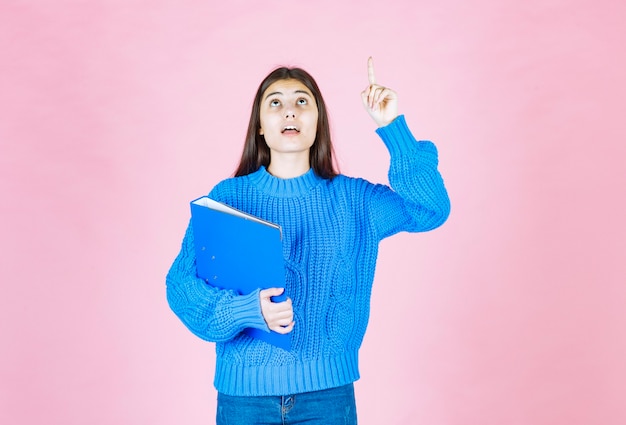 niña sosteniendo una carpeta azul y apuntando hacia arriba en rosa.