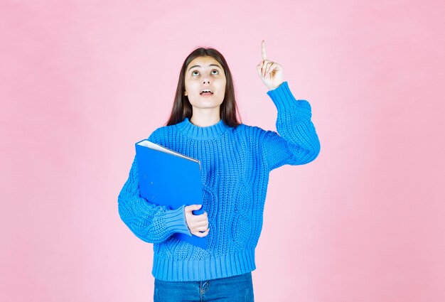 niña sosteniendo una carpeta azul y apuntando hacia arriba en rosa.