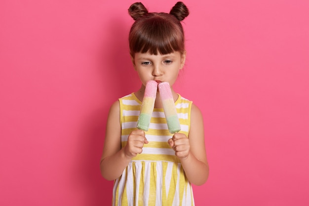 Niña sosteniendo y comiendo dos helados, mirando hacia abajo, pensativa, se ve pensativa, posando aislada sobre la pared de rosas, disfrutando de un sabroso sorbete de verano.