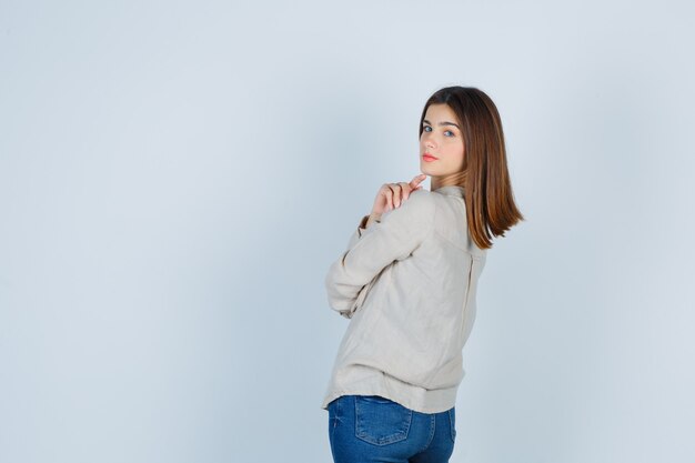 Niña sosteniendo el dedo índice debajo de la barbilla, mirando por encima del hombro con camisa beige, jeans y aspecto encantador. vista frontal.