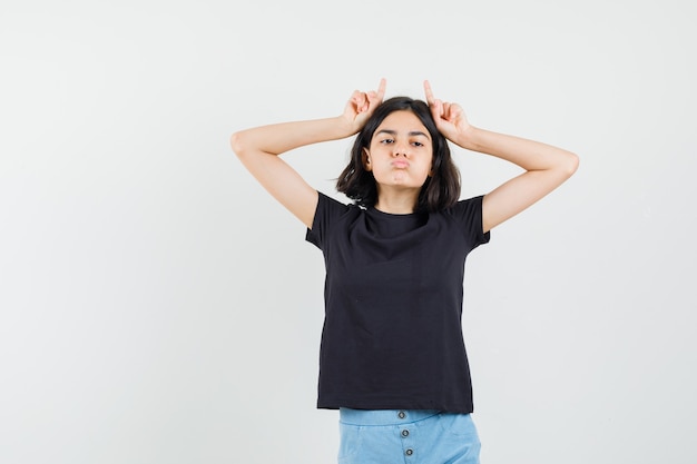 Niña sosteniendo los dedos sobre la cabeza como cuernos de toro en camiseta negra, pantalones cortos y mirando divertido, vista frontal.