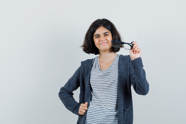 Niña sosteniendo gafas en camiseta, chaqueta y mirando optimista,