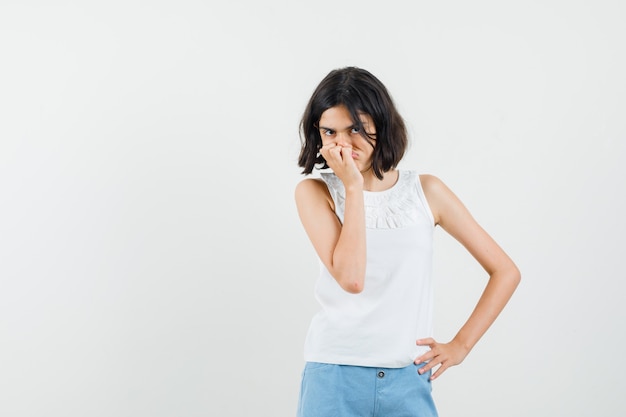 Foto gratuita niña sosteniendo la mano en la boca en blusa blanca, pantalones cortos y mirando resentido, vista frontal.