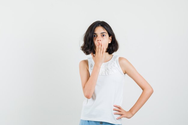 Niña sosteniendo la mano en la boca en blusa blanca, pantalones cortos y mirando sorprendido, vista frontal.