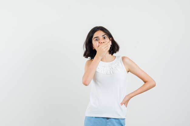 Niña sosteniendo la mano en la boca en blusa blanca, pantalones cortos y mirando sorprendido, vista frontal.