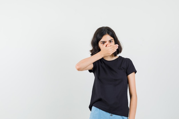 Niña sosteniendo la mano en la boca en camiseta negra, pantalones cortos y mirando asustada. vista frontal.