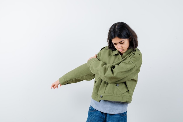 Niña sosteniendo la mano en el brazo en abrigo, camiseta, jeans y mirando pensativo. vista frontal.