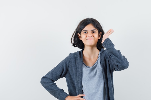 Niña sosteniendo la mano cerca de la oreja en camiseta, chaqueta y mirando alegre. vista frontal.