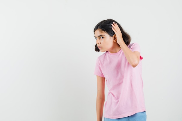 Foto gratuita niña sosteniendo la mano cerca de la oreja en camiseta rosa, pantalones cortos y con curiosidad. vista frontal.