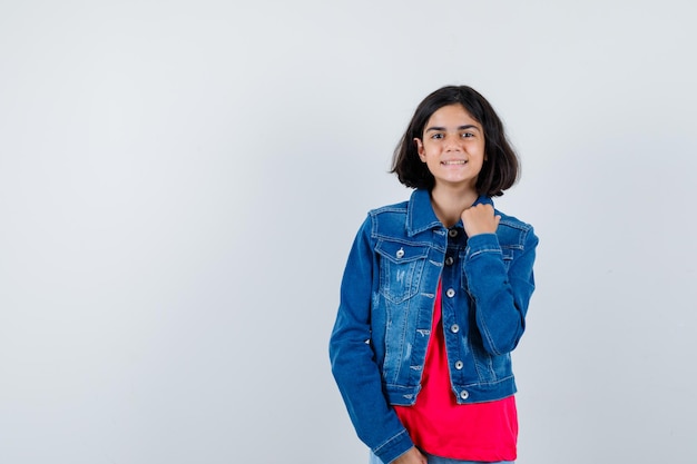 Niña sosteniendo la mano en la chaqueta en camiseta roja y chaqueta de mezclilla y mirando feliz, vista frontal.