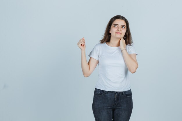 Niña sosteniendo la mano en el cuello mientras mira a otro lado en camiseta, jeans y mirando pensativo. vista frontal.