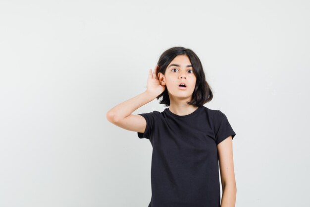 Niña sosteniendo la mano detrás de la oreja en camiseta negra y mirando curioso, vista frontal.