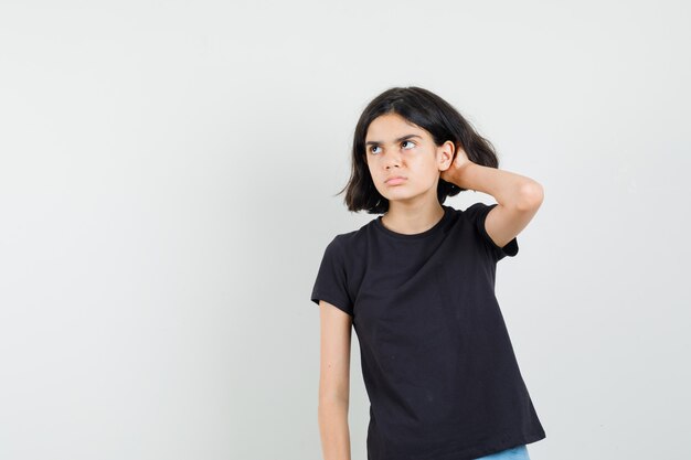 Niña sosteniendo la mano detrás de la oreja en camiseta negra y mirando curioso, vista frontal.