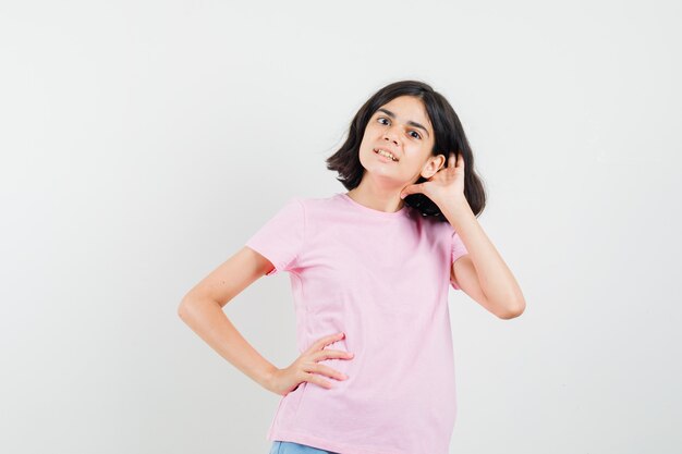 Niña sosteniendo la mano detrás de la oreja en camiseta rosa, pantalones cortos y con curiosidad. vista frontal.