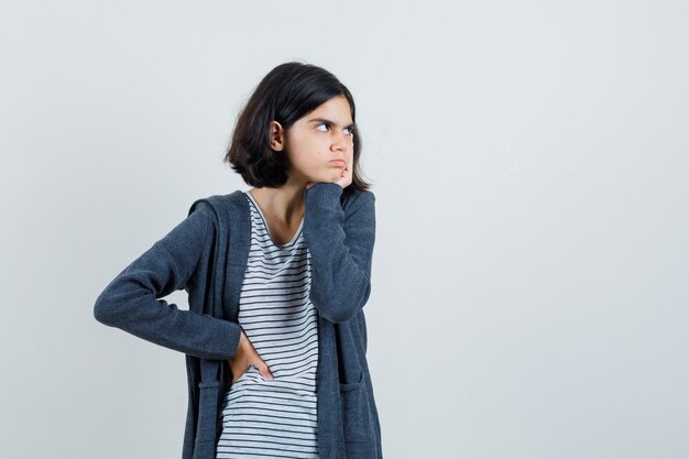 Niña sosteniendo la mano en la mejilla en camiseta, chaqueta y mirando triste.