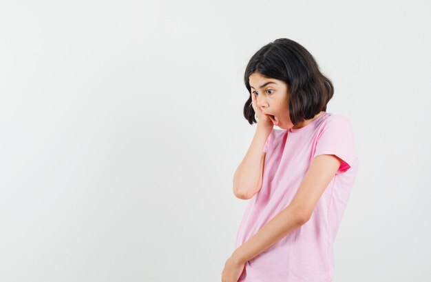 Niña sosteniendo la mano en la mejilla en camiseta rosa y mirando sorprendido. vista frontal.