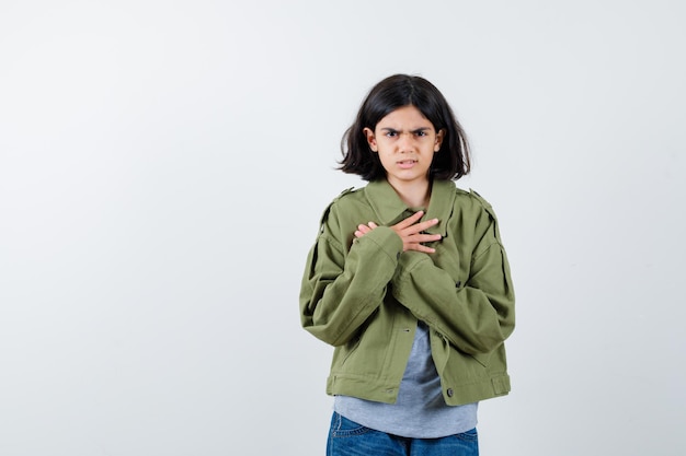 Niña sosteniendo las manos cruzadas sobre el pecho en abrigo, camiseta, jeans y mirando nostálgico, vista frontal.