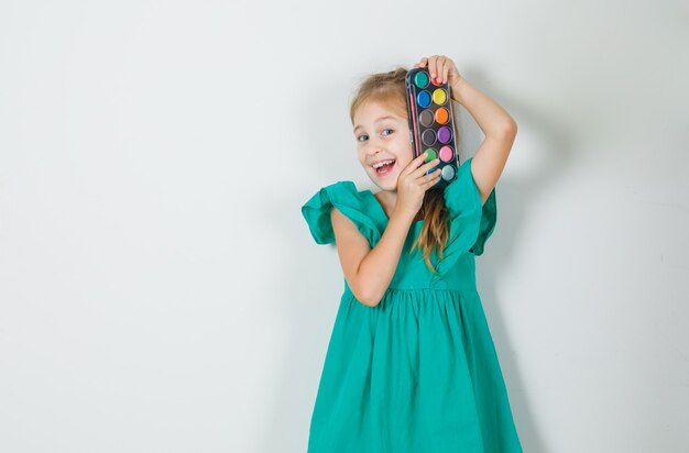 Niña sosteniendo pinturas de acuarela con pincel en vestido verde y mirando alegre