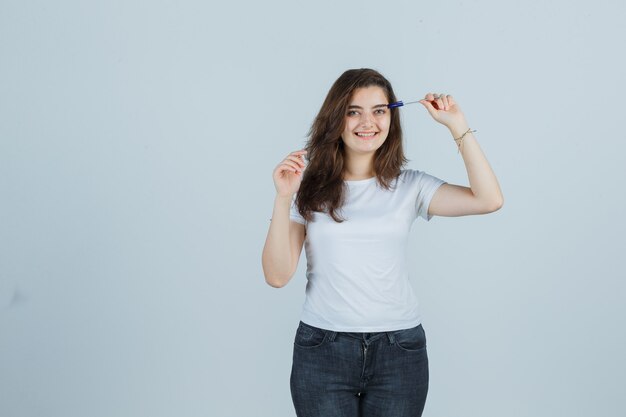 Niña sosteniendo la pluma en la cabeza en camiseta, jeans y mirando alegre. vista frontal.