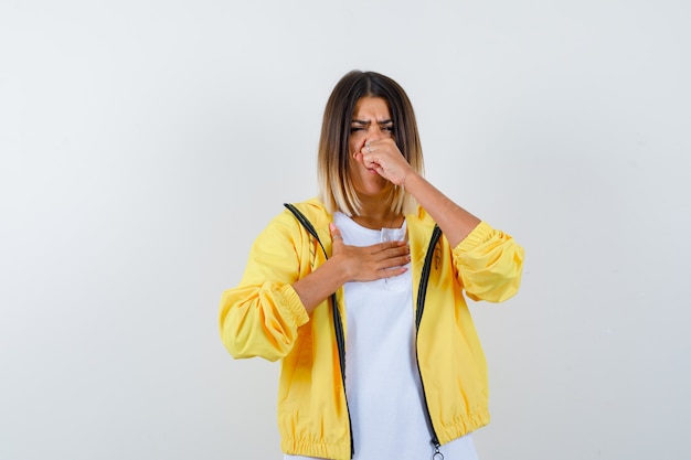 Foto gratuita niña sosteniendo el puño cerca de la boca, estornudando en camiseta blanca, chaqueta amarilla y mirando exhausto, vista frontal.