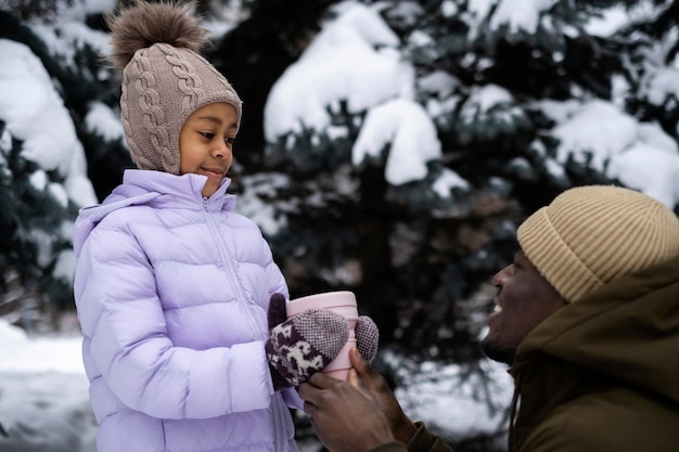 Niña sosteniendo una taza de bebida caliente mientras sale en un día de invierno con su padre