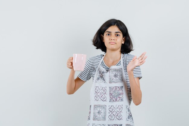 Niña sosteniendo una taza de bebida en camiseta, delantal y mirando confundido.