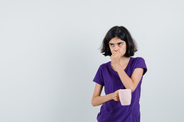 Niña sosteniendo una taza de bebida en camiseta y mirando pensativa.