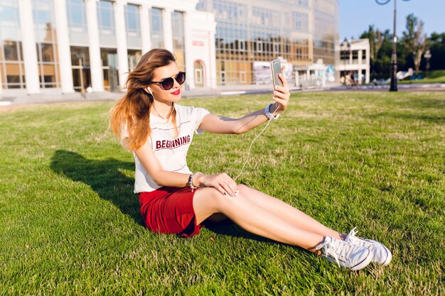 Niña sosteniendo un teléfono inteligente y escuchando música se sienta en la hierba verde en el parque de la ciudad