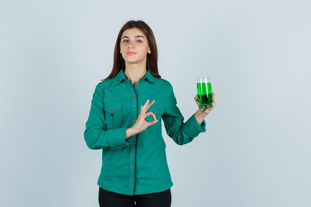 Niña sosteniendo un vaso de líquido verde, mostrando el signo de ok en blusa verde, pantalón negro y mirando alegre, vista frontal.