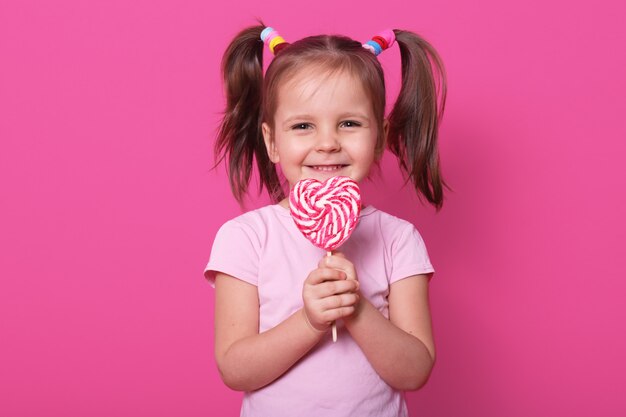 La niña sostiene una enorme paleta de rayas en forma de corazón, se ve feliz y emocionada, usa una camiseta rosa, se encuentra sonriente aislado en rosa. A la niña le gustan los dulces.