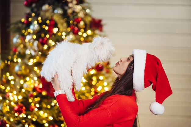 Una niña sostiene un perro pequeño en sus manos en el árbol de navidad