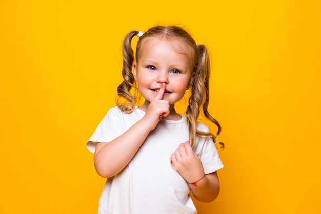 Niña con su dedo sobre la boca diciendo Shh aislado sobre pared amarilla