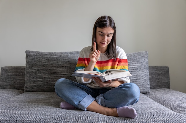 Foto gratuita una niña con un suéter de colores brillantes lee un libro en el sofá de casa.