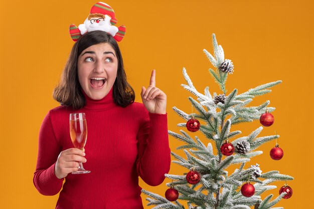 Niña en suéter de navidad con diadema divertida sosteniendo una copa de champán feliz y sorprendido junto a un árbol de navidad sobre fondo naranja