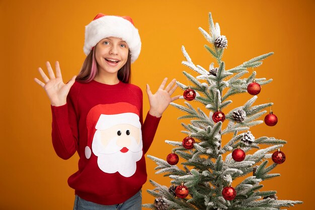 Niña en suéter de navidad y gorro de Papá Noel feliz y sorprendido sonriendo alegremente de pie junto a un árbol de navidad sobre fondo naranja