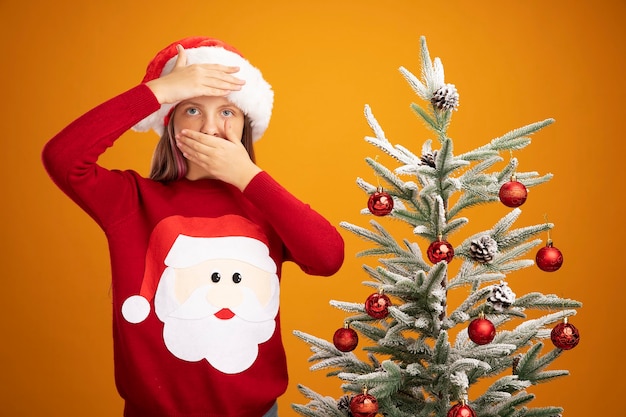 Niña en suéter de Navidad y gorro de Papá Noel mirando a la cámara con la mano en la frente cubriendo la boca con la otra mano de pie junto a un árbol de Navidad sobre fondo naranja