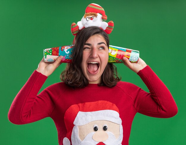 Foto gratuita niña en suéter de navidad con gorro de papá noel sosteniendo vasos de papel de colores cerca de sus oídos, feliz y emocionado de pie sobre la pared verde