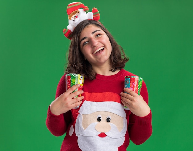 Foto gratuita niña de suéter de navidad con gorro de papá noel sosteniendo vasos de papel de colores mirando a la cámara sonriendo feliz y alegre de pie sobre fondo verde