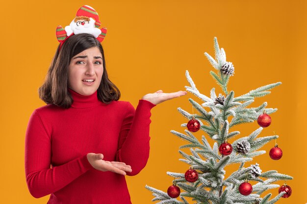 Niña de suéter de Navidad vistiendo una diadema divertida junto a un árbol de Navidad sobre fondo naranja