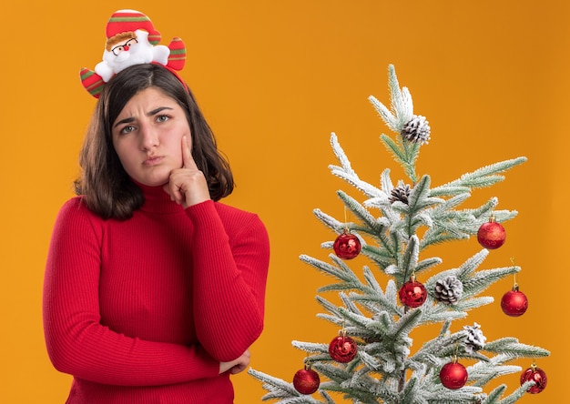 Niña de suéter de Navidad vistiendo una diadema divertida junto a un árbol de Navidad sobre fondo naranja