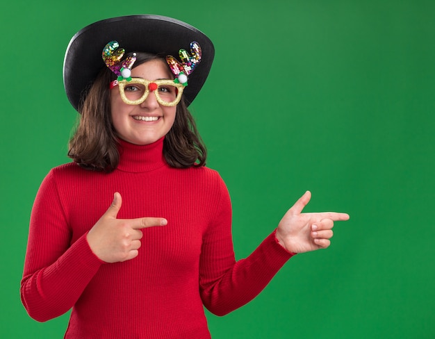 Niña de suéter rojo con gafas divertidas y sombrero negro mirando a la cámara sonriendo