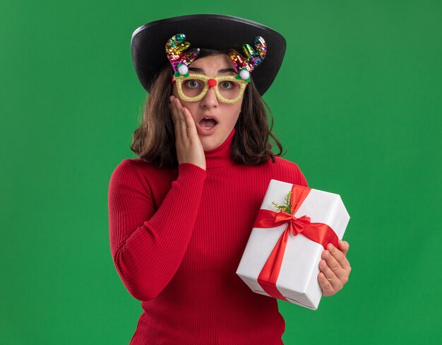 Niña de suéter rojo con gafas divertidas y sombrero negro sosteniendo un presente sorprendido y feliz de pie sobre la pared verde