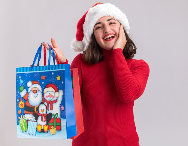 Foto gratuita niña de suéter rojo y gorro de papá noel con bolsas de papel de colores con regalos de navidad mirando a la cámara con cara feliz de pie sobre fondo blanco.
