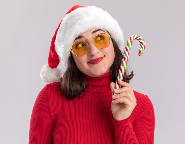 Niña de suéter rojo y gorro de Papá Noel con gafas sosteniendo bastón de caramelo mirando hacia arriba con una sonrisa en la cara de pie sobre fondo blanco.
