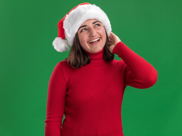 Niña en suéter rojo y gorro de Papá Noel mirando hacia arriba con una sonrisa en la cara feliz y alegre de pie sobre la pared verde