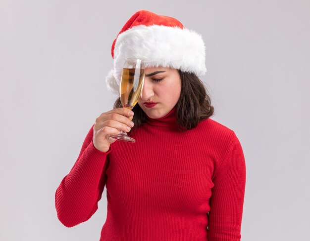 Niña de suéter rojo y gorro de Papá Noel sosteniendo una copa de champán con aspecto cansado y aburrido de pie sobre fondo blanco.