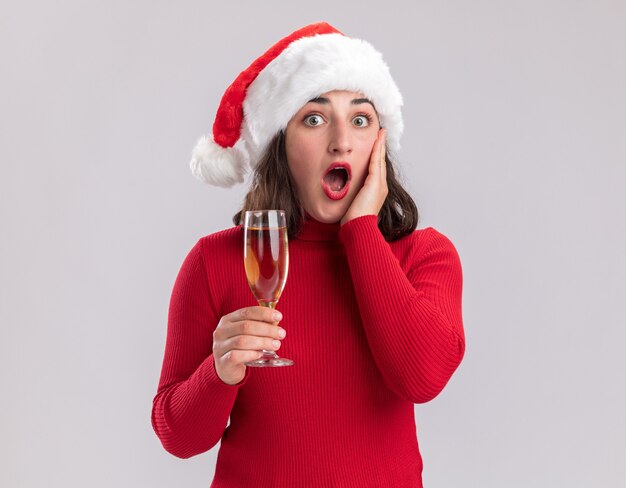 Niña de suéter rojo y gorro de Papá Noel sosteniendo una copa de champán mirando a la cámara sorprendida de pie sobre fondo blanco.