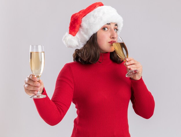 Niña de suéter rojo y gorro de Papá Noel sosteniendo dos copas de champán bebiendo feliz y positivo de pie sobre fondo blanco.