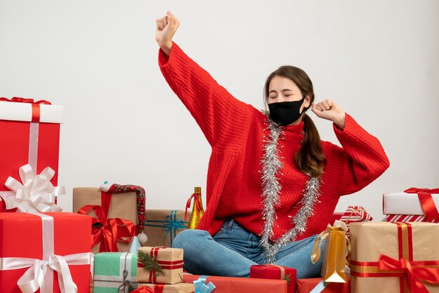 Niña con suéter rojo y máscara negra abriendo las manos sentados alrededor de regalos en blanco