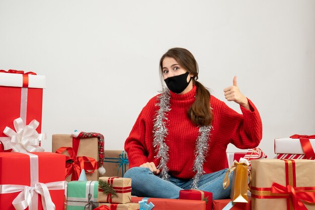 Niña con suéter rojo y máscara negra haciendo pulgar hacia arriba firmar sentados alrededor de regalos en blanco
